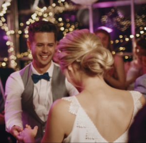 Man in a gray vest and blue bow tie dancing with a woman in a white lace dress at a party with warm string lights in the background.