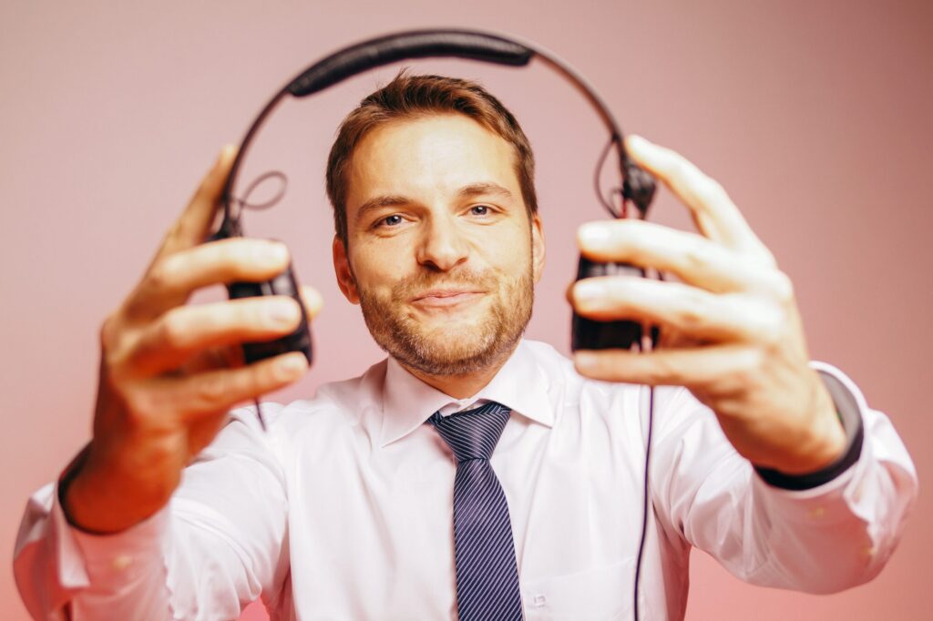 Man in a white shirt and striped tie holds over-ear headphones toward the camera, smiling against a pink background.
